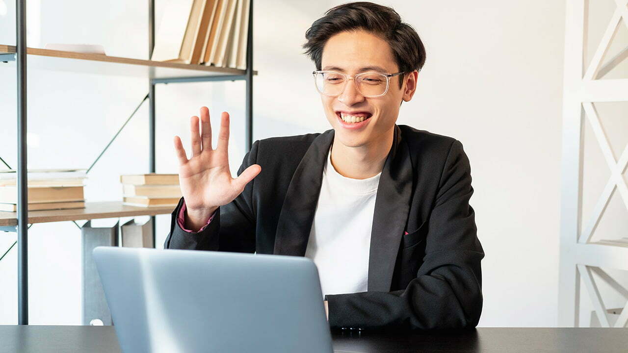 Young man with glasses waving and smiling at his laptop during a video call