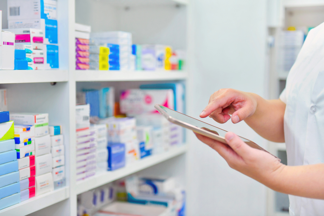 Pharmacist in a white coat using a tablet in front of shelves full of medicine boxes