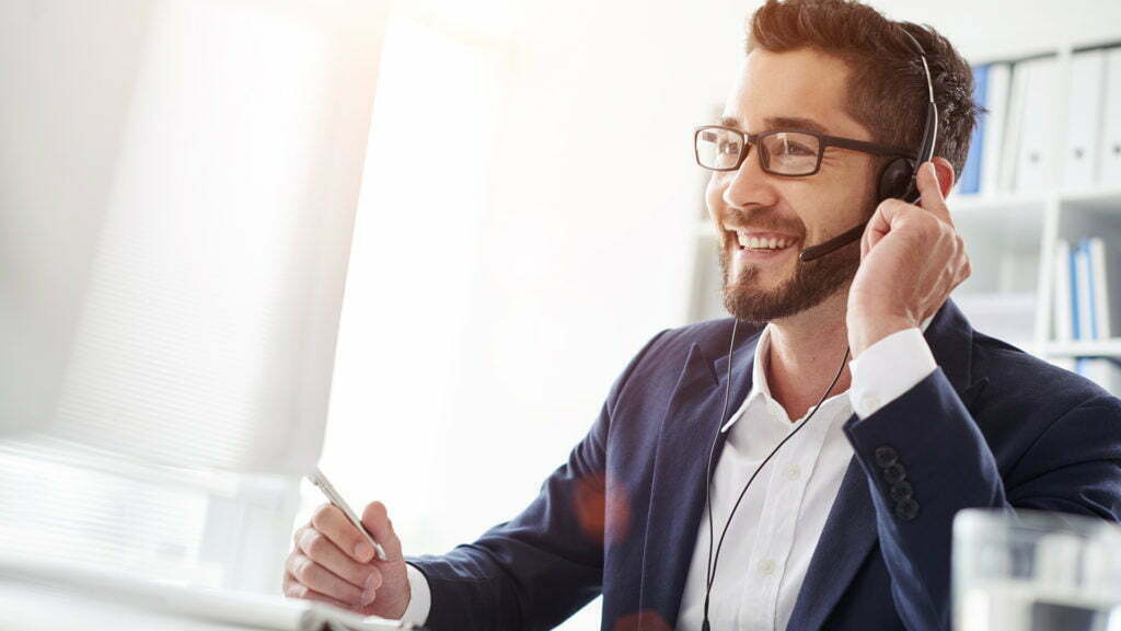 Smiling man with glasses and headset in a suit, sitting at a computer in an office