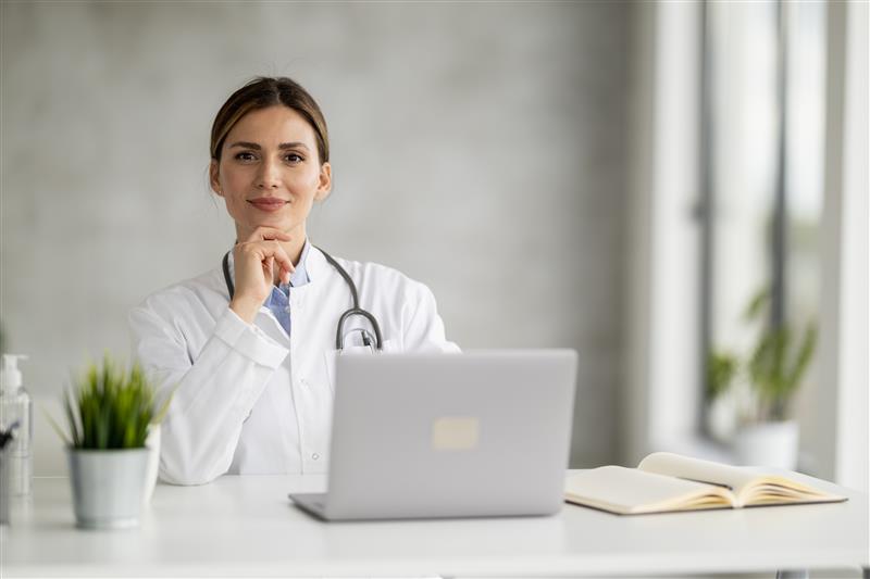 Doctor in a white coat with a stethoscope sitting smiling behind a laptop at a desk