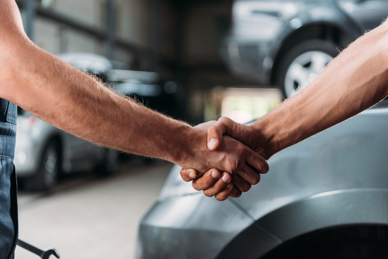 Two people shaking hands in a garage, with cars in the background and a spanner in hand