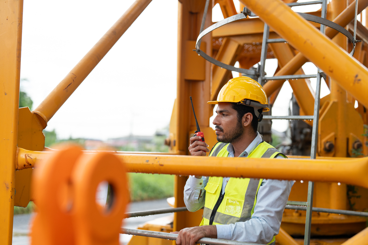 Construction worker in a helmet and safety vest talking into a two-way radio on a yellow crane