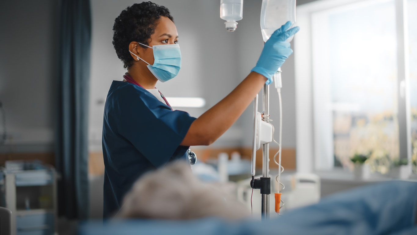 Nurse wearing a face mask and gloves checking an IV bag next to a patient’s bed