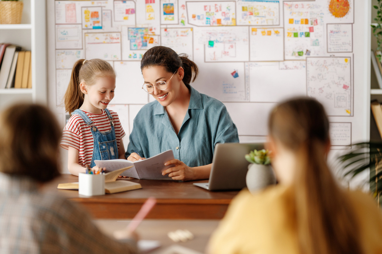 Teacher helping a girl with a notebook in a classroom, with other pupils in the foreground