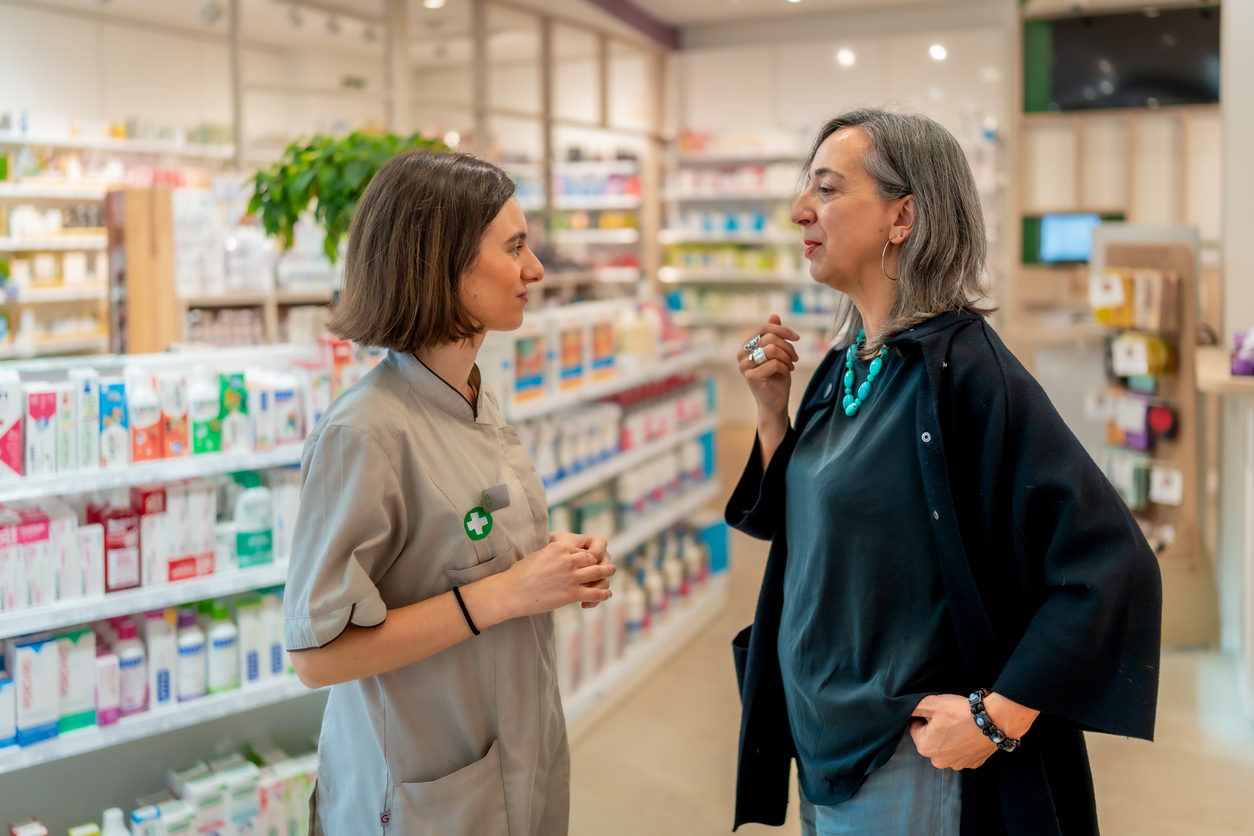Pharmacy assistant talking to an older woman in a pharmacy with well-stocked shelves