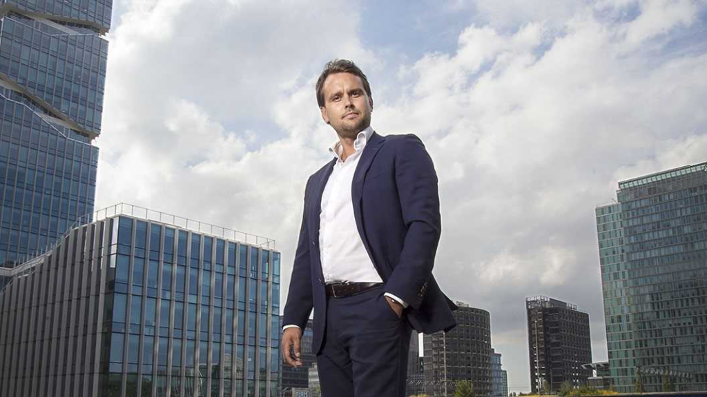 Man in a dark suit and white shirt standing against a cloudy sky and glass office towers