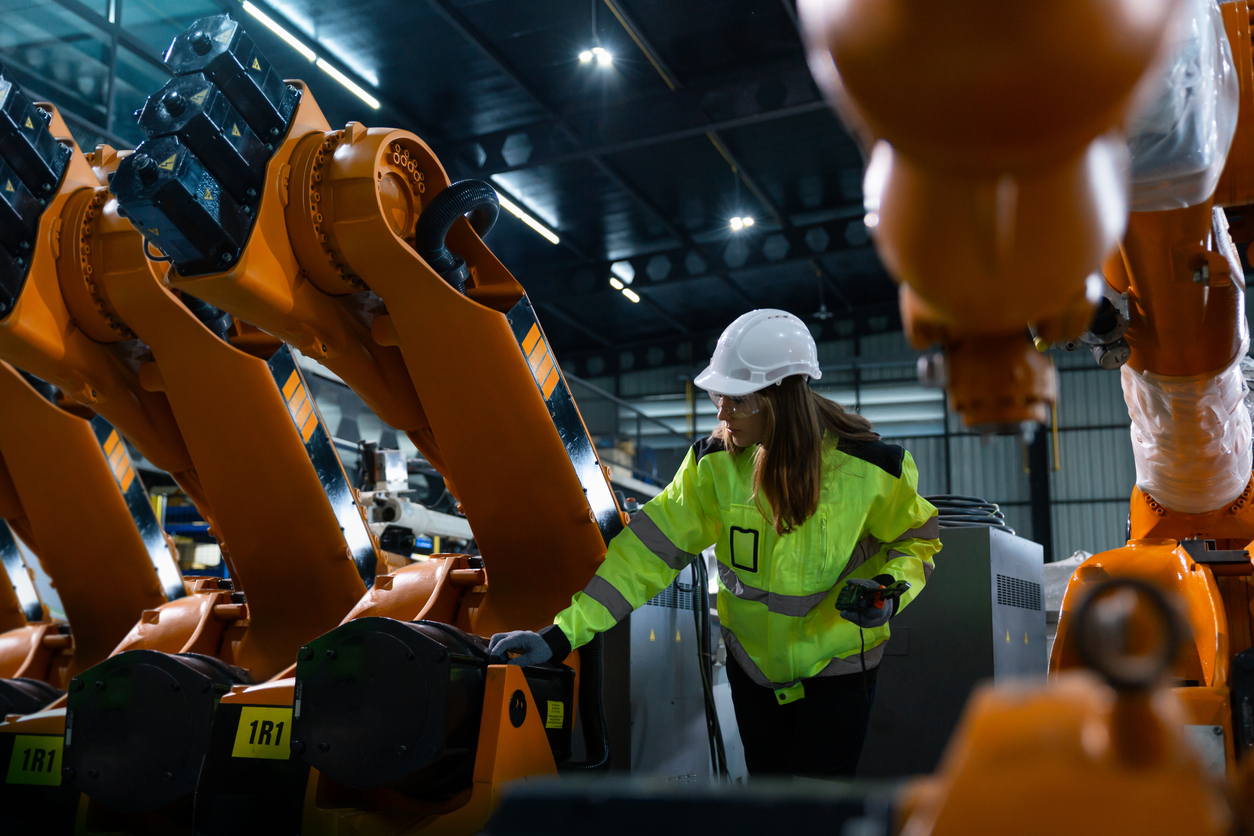 Woman in a helmet and safety jacket inspecting orange robot arms in a factory hall