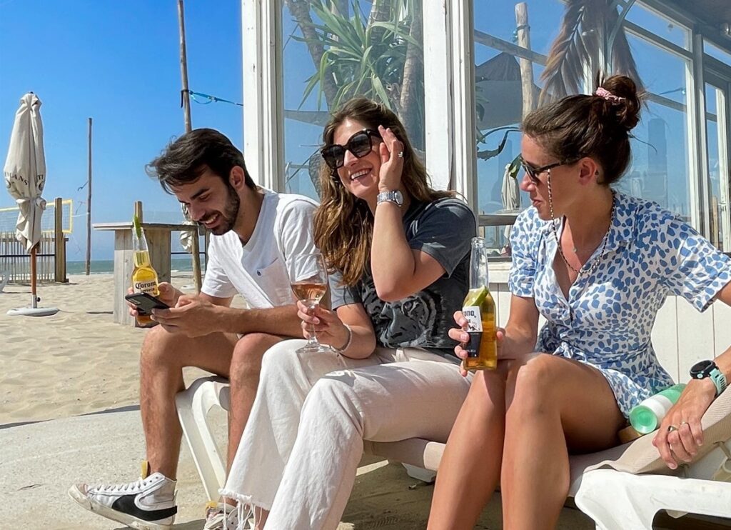 Three young people laughing on a beach terrace with drinks and a phone