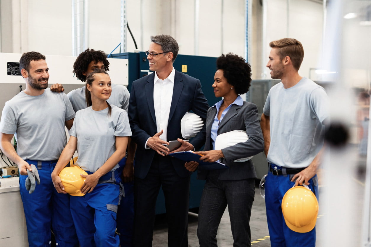 Factory workers in helmets laughing together with a man in a suit and a woman with a clipboard