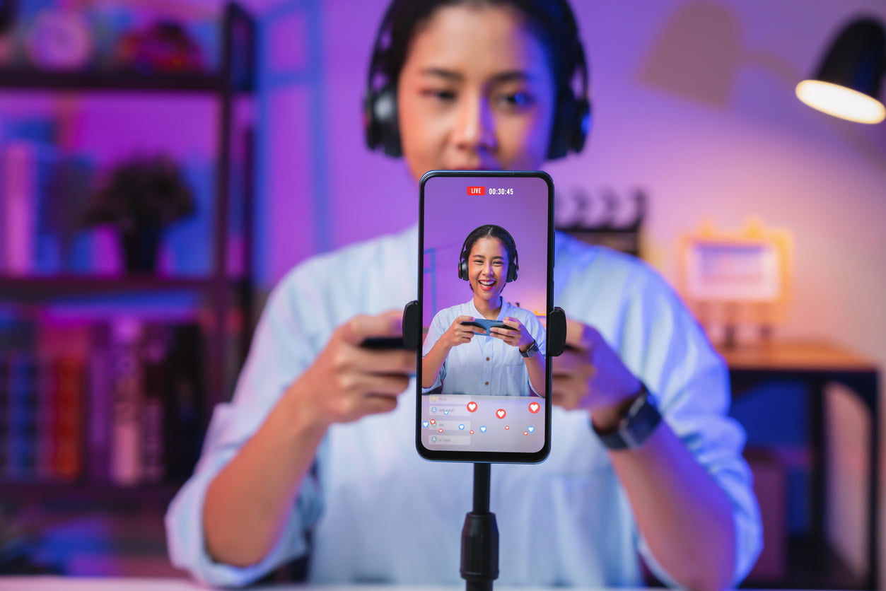 Woman wearing headphones live-streaming via a smartphone on a tripod, in purple-blue light