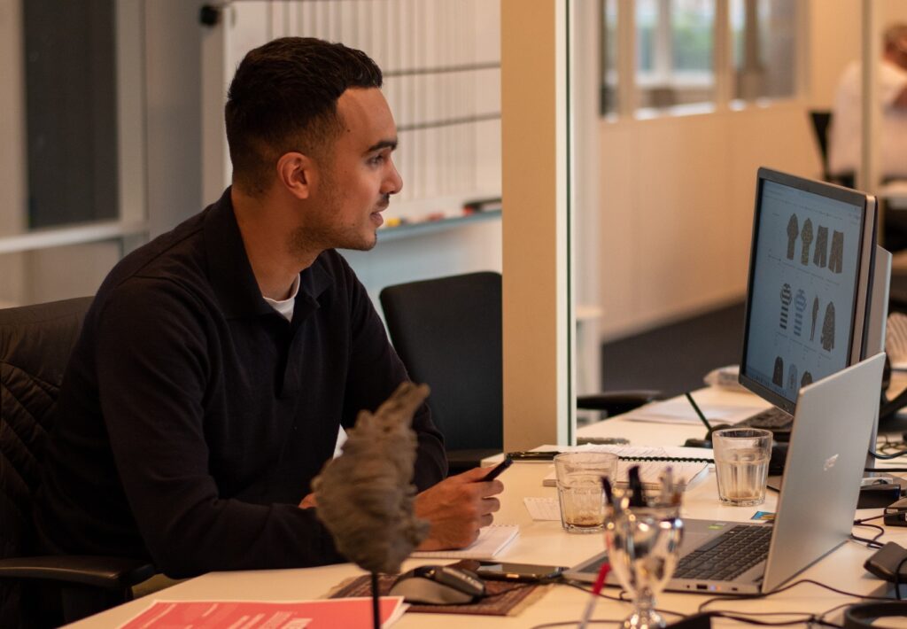 Man in a dark shirt looking sideways at his desk, with laptop and monitor in an open-plan office
