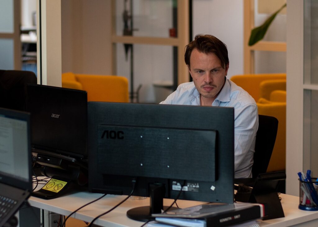 Man in a light blue shirt working at a monitor in an open-plan office with yellow chairs
