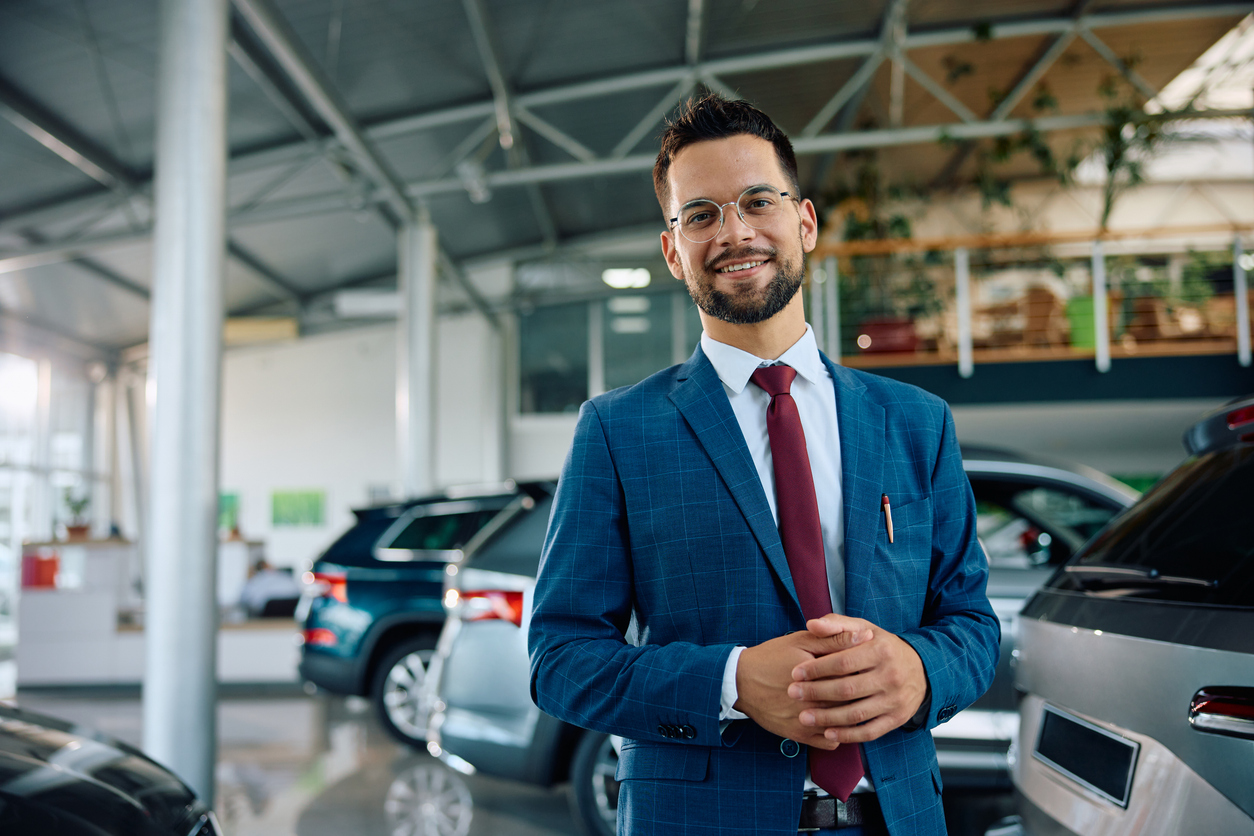 Smiling car salesman in a blue suit standing in a showroom between cars