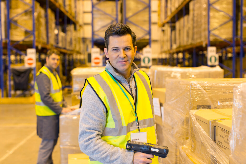 Warehouse operative in a high-visibility vest with a handheld scanner between racks full of pallets, colleague in the background