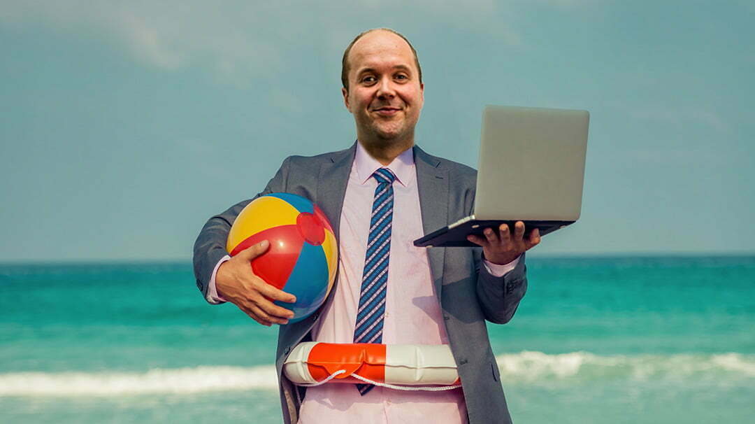 Man in a suit on the beach holding a laptop and a beach ball, wearing a swim ring