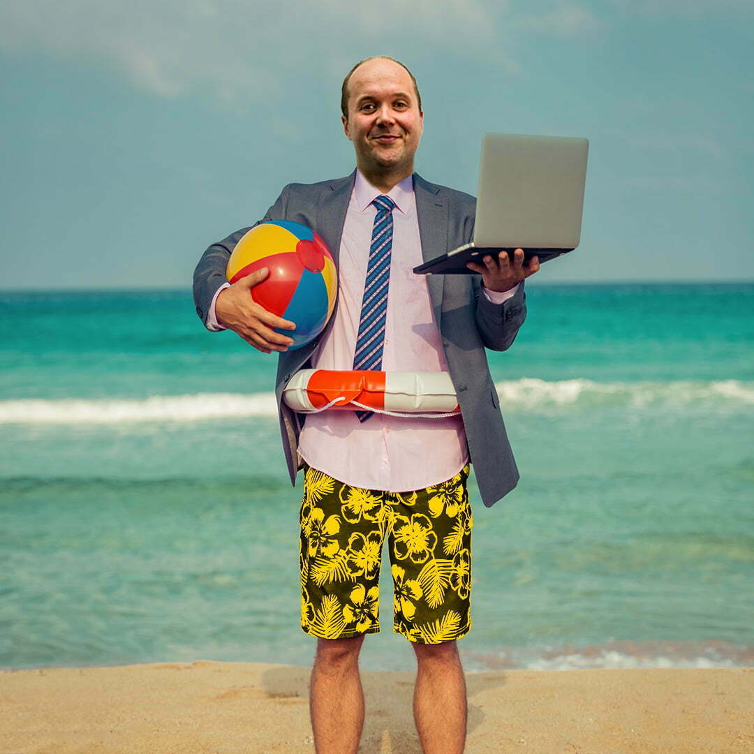 Man in a suit and swimming trunks standing on the beach with a laptop, beach ball and swim ring