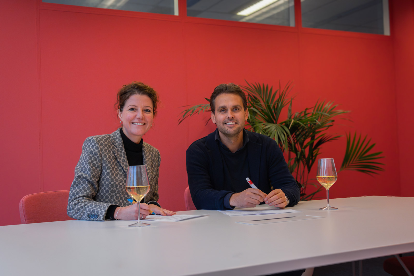 Man and woman sitting at a table in front of a red wall with wine glasses, signing documents