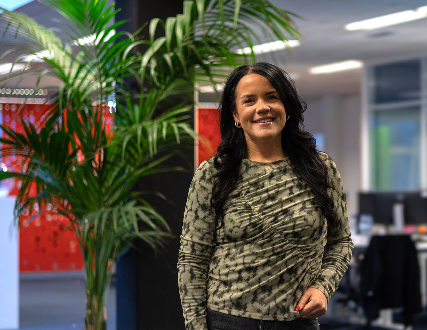 Smiling woman with long dark hair standing next to a palm plant in an office