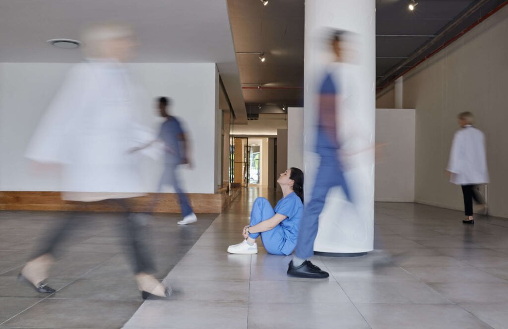 Nurse in blue scrubs sitting on the floor of a corridor while colleagues walk past in a blur