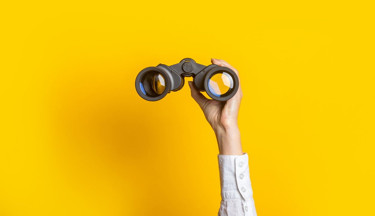 Hand holding up a pair of binoculars against a yellow background