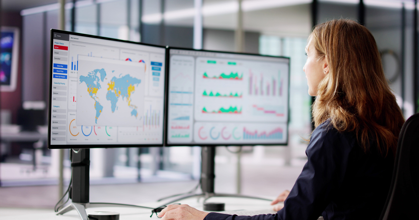 Woman looking at two monitors showing charts and a world map