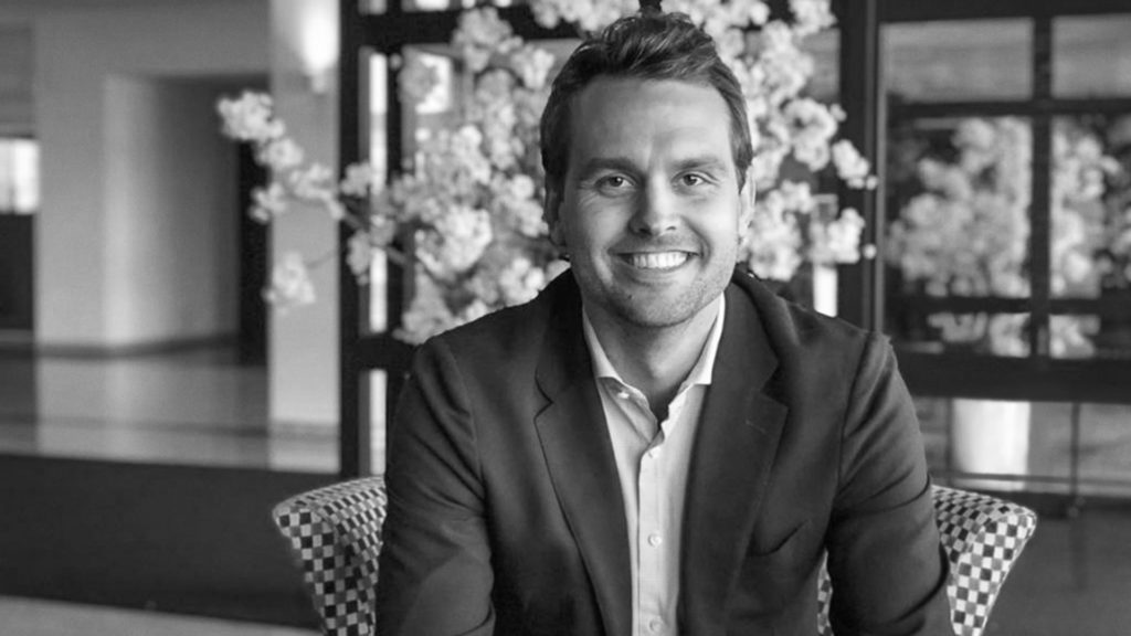Black and white portrait of a smiling man in a blazer, sitting in a hall with blossom branches