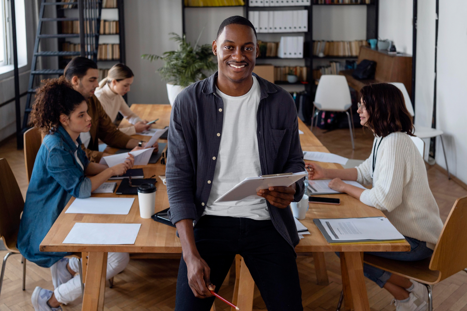 Smiling man with a clipboard standing in front of a table where colleagues are working