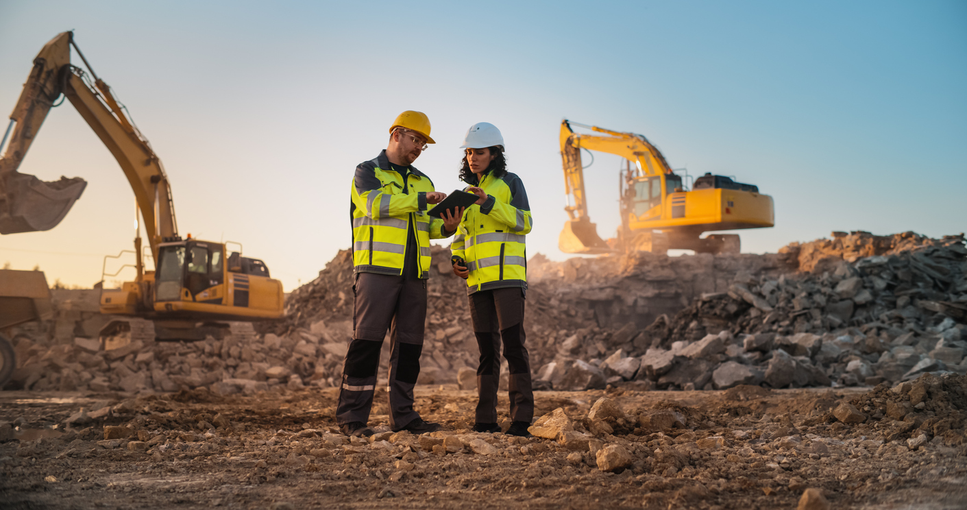 Two construction workers in yellow vests looking at a tablet on a building site with excavators
