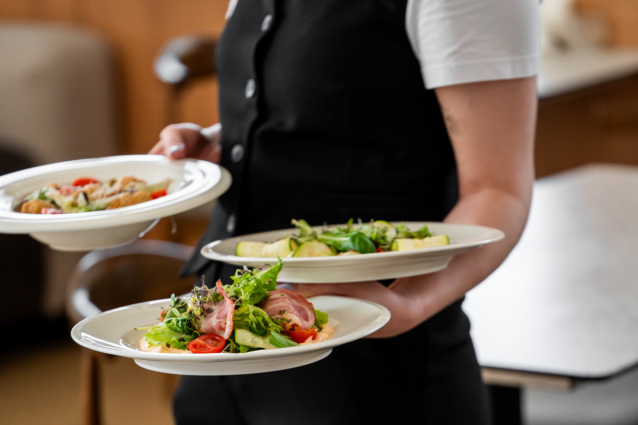 Staff member in a black waistcoat carrying three plates of fresh salads