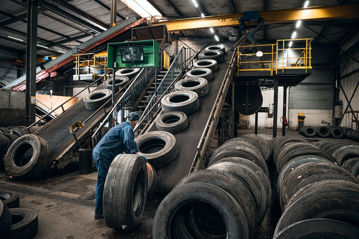 Worker in blue overalls rolling a truck tyre towards conveyor belts in a tyre recycling hall