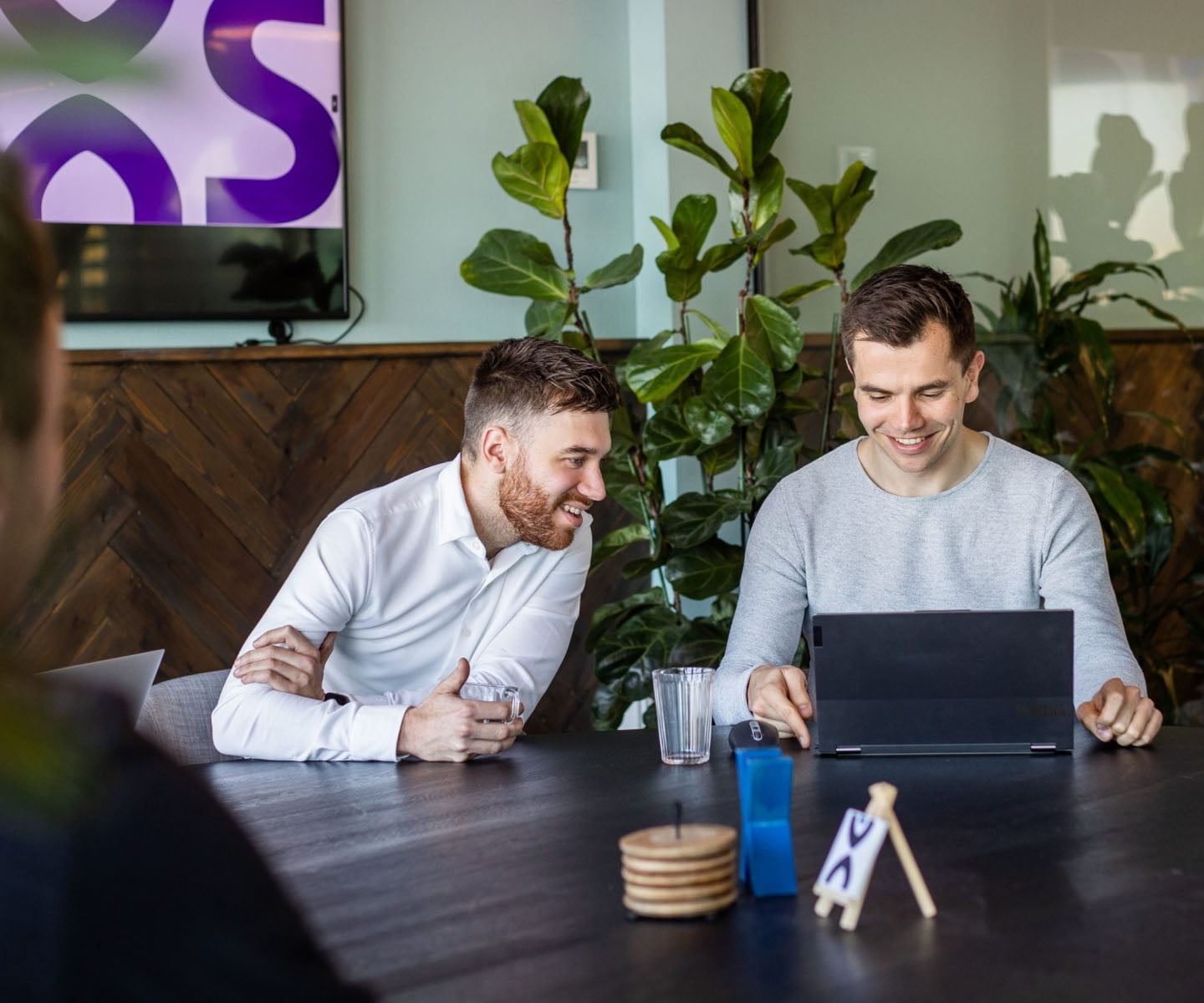 Two smiling men looking at a laptop together at a table with plants, a purple logo on the screen behind them