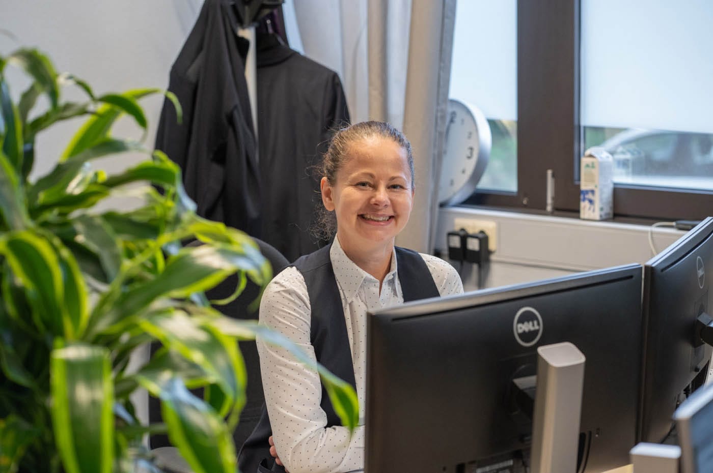 Smiling employee in a waistcoat sitting at a computer screen in an office, with a plant in the foreground