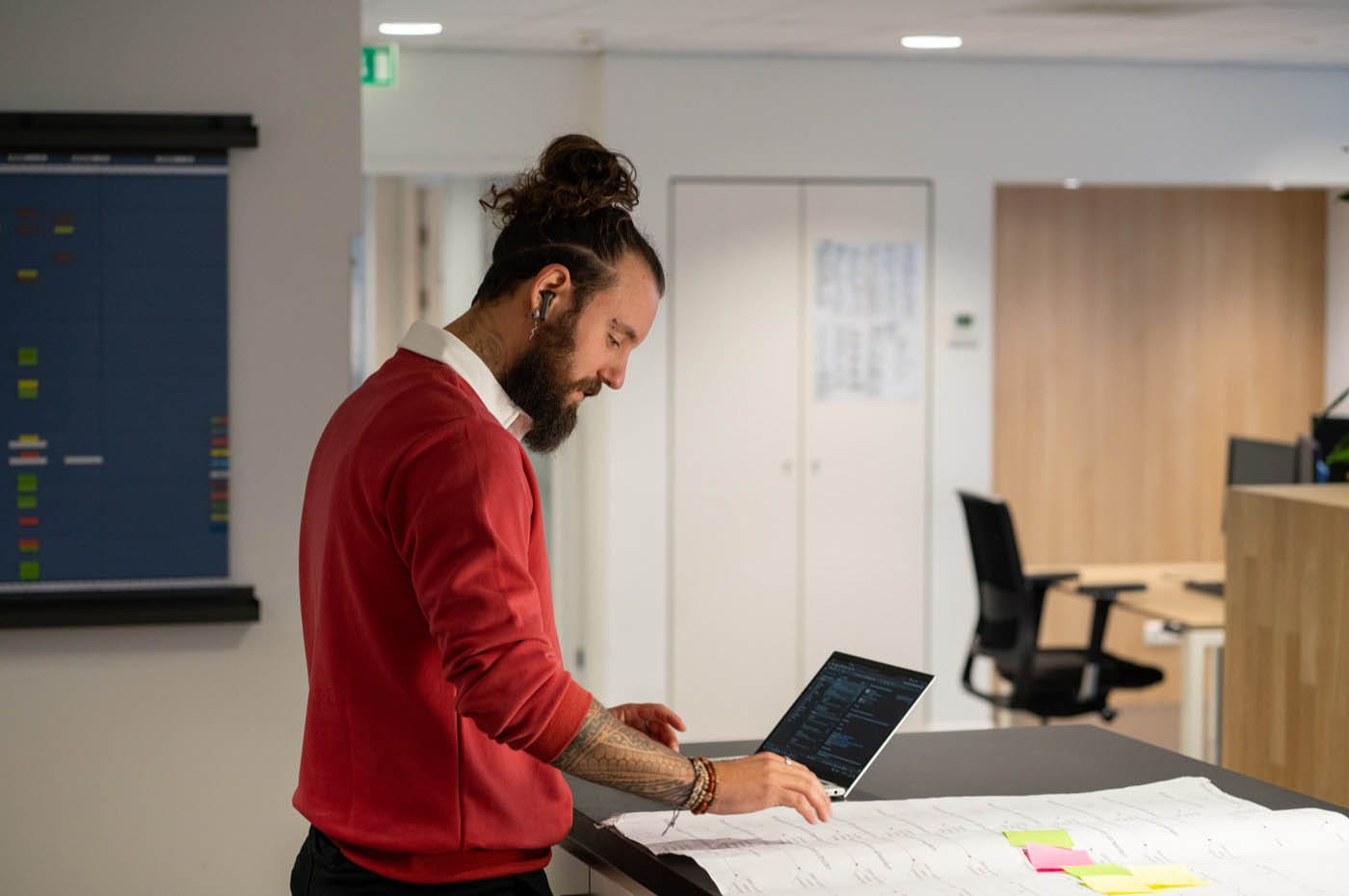 Man in a red sweater standing at a laptop next to large drawings with sticky notes, a planning board behind him