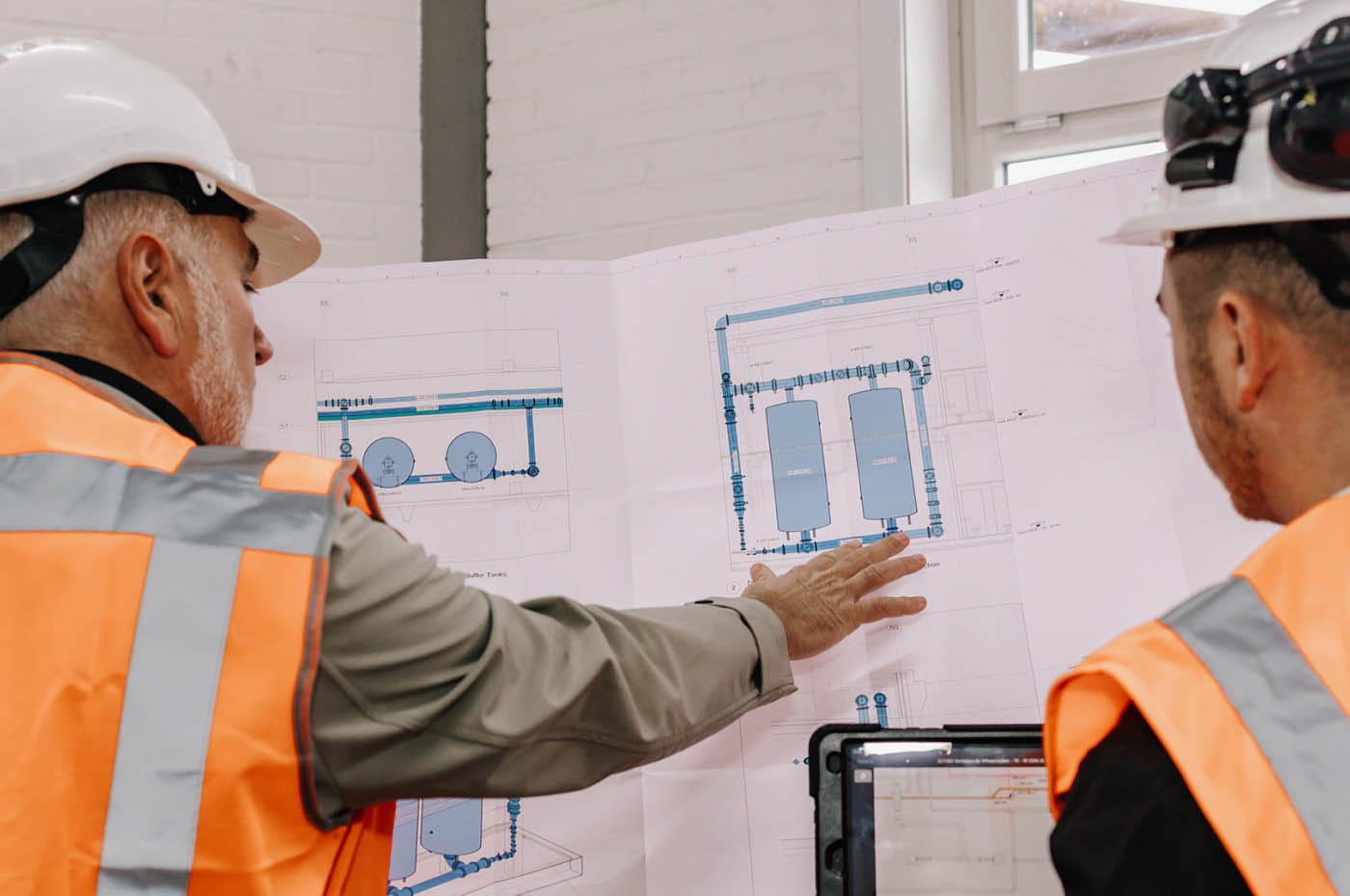 Two men in white hard hats and orange high-vis vests discussing a construction drawing with piping diagrams
