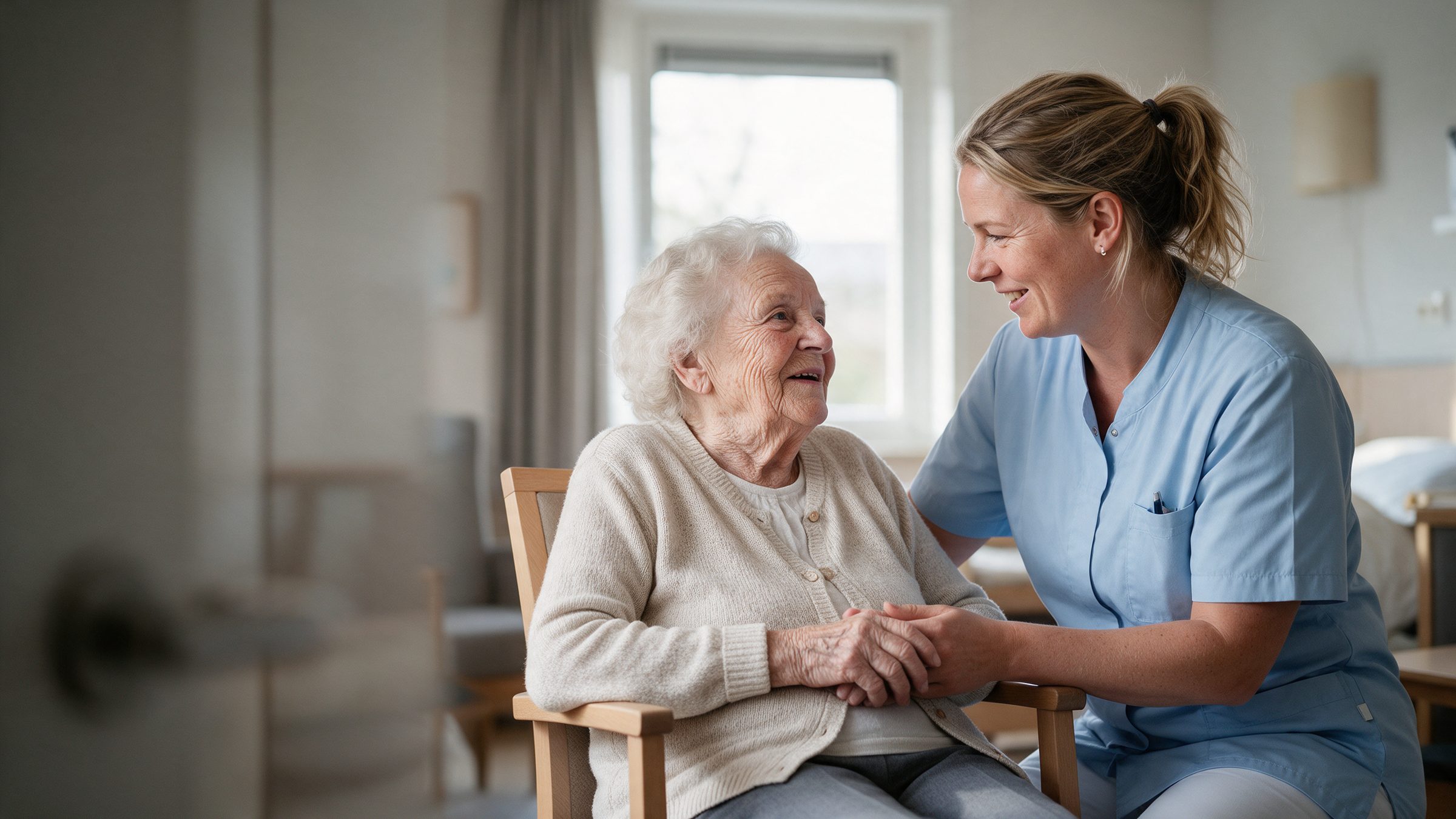 Carer in a light blue uniform holding the hands of a smiling elderly woman