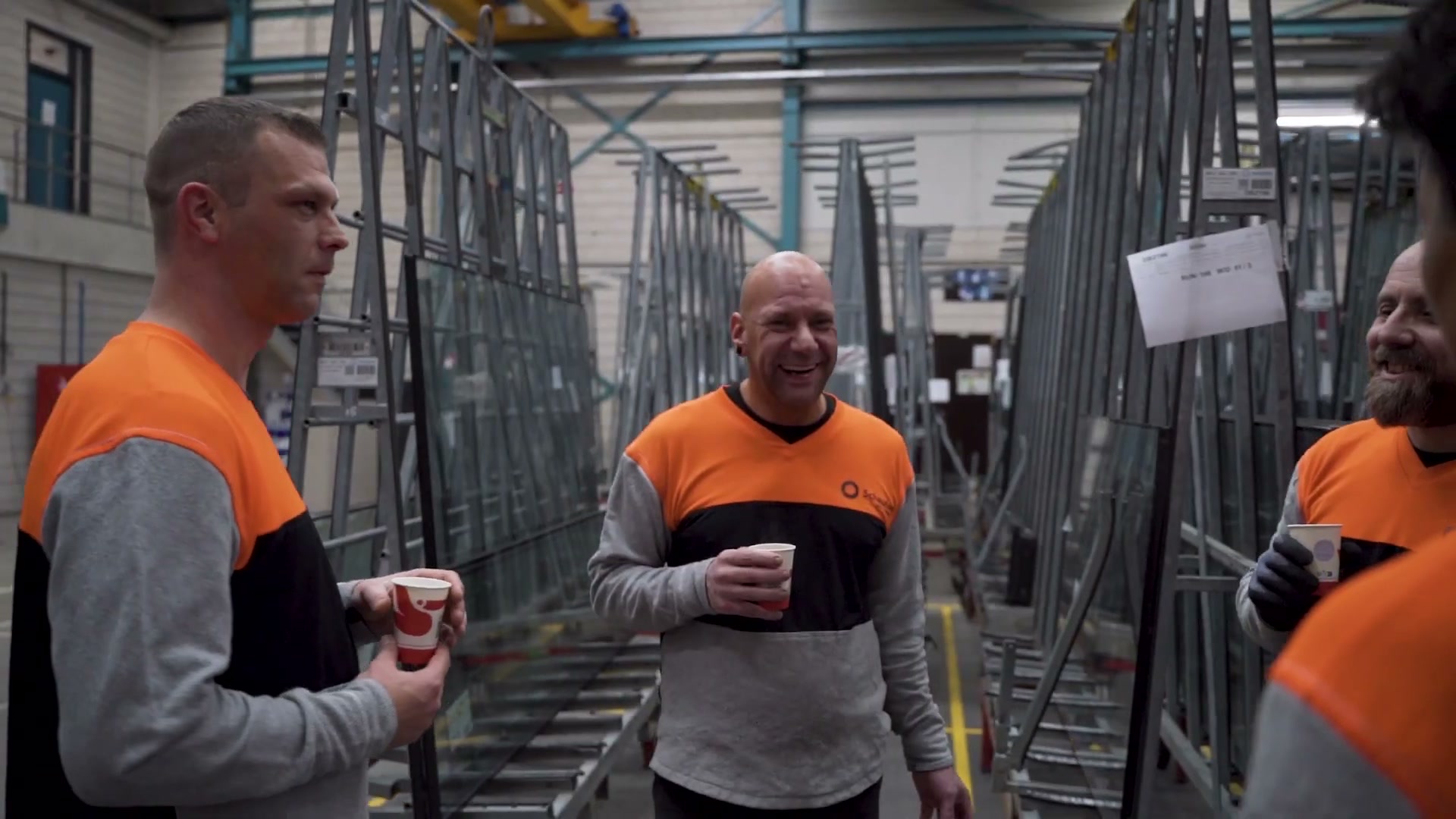 Two Scheuten Glas employees laughing over a cup of coffee between the glass racks in the factory