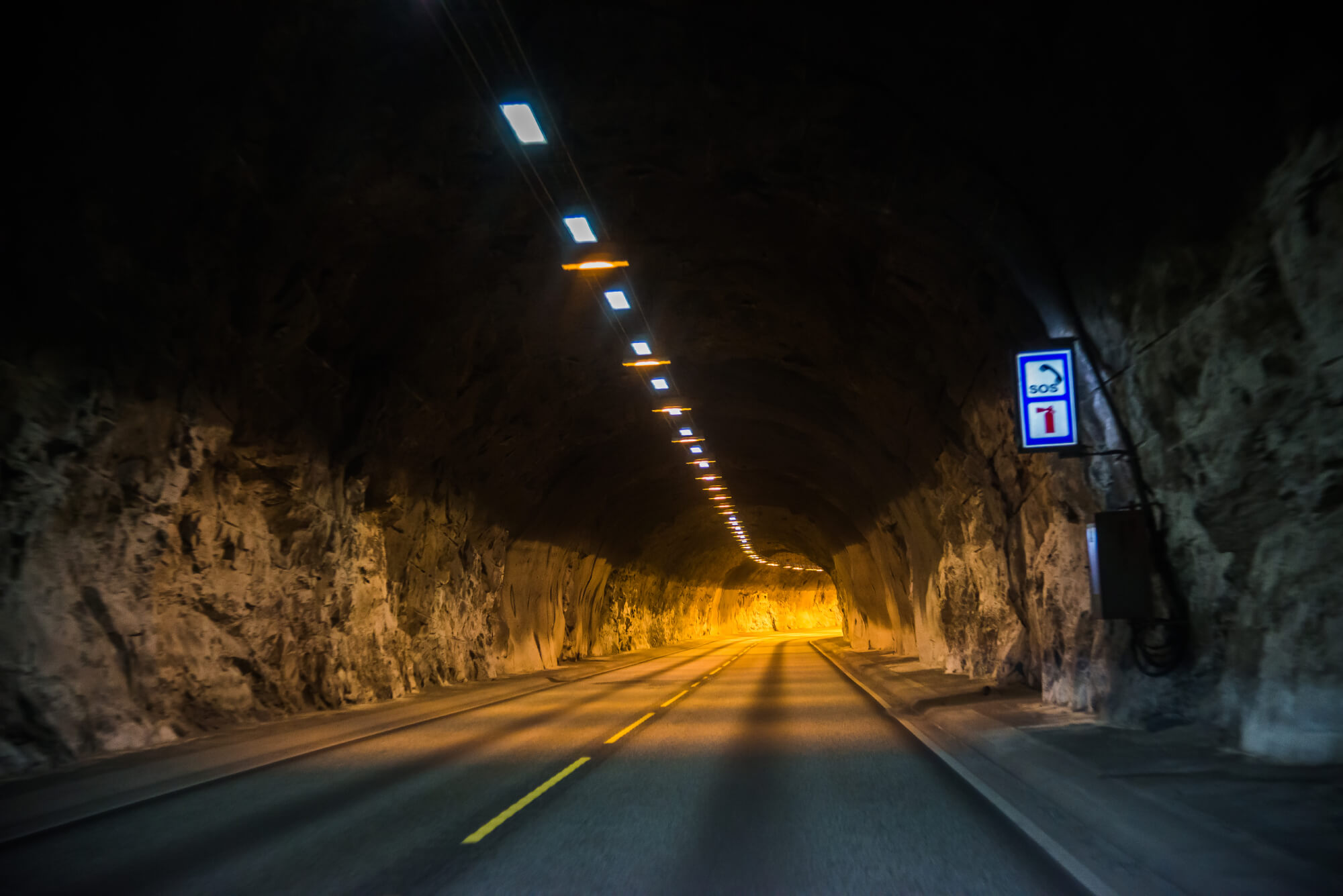 Empty two-lane road inside a dimly lit rock tunnel with overhead lights and an SOS sign on the right wall.