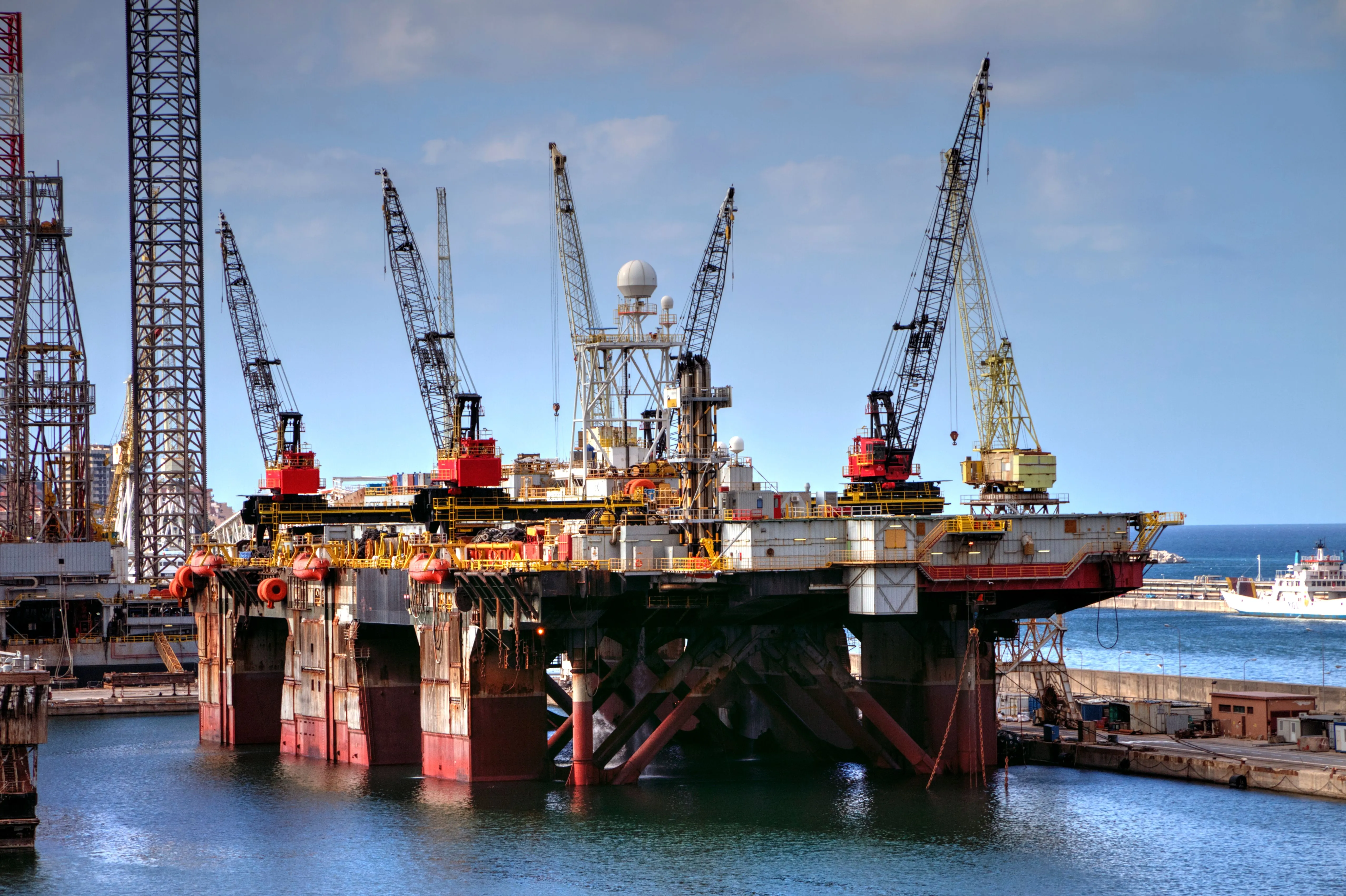 Large offshore oil drilling platform docked in a harbor with multiple cranes and equipment on the deck, surrounded by calm water and a clear blue sky.