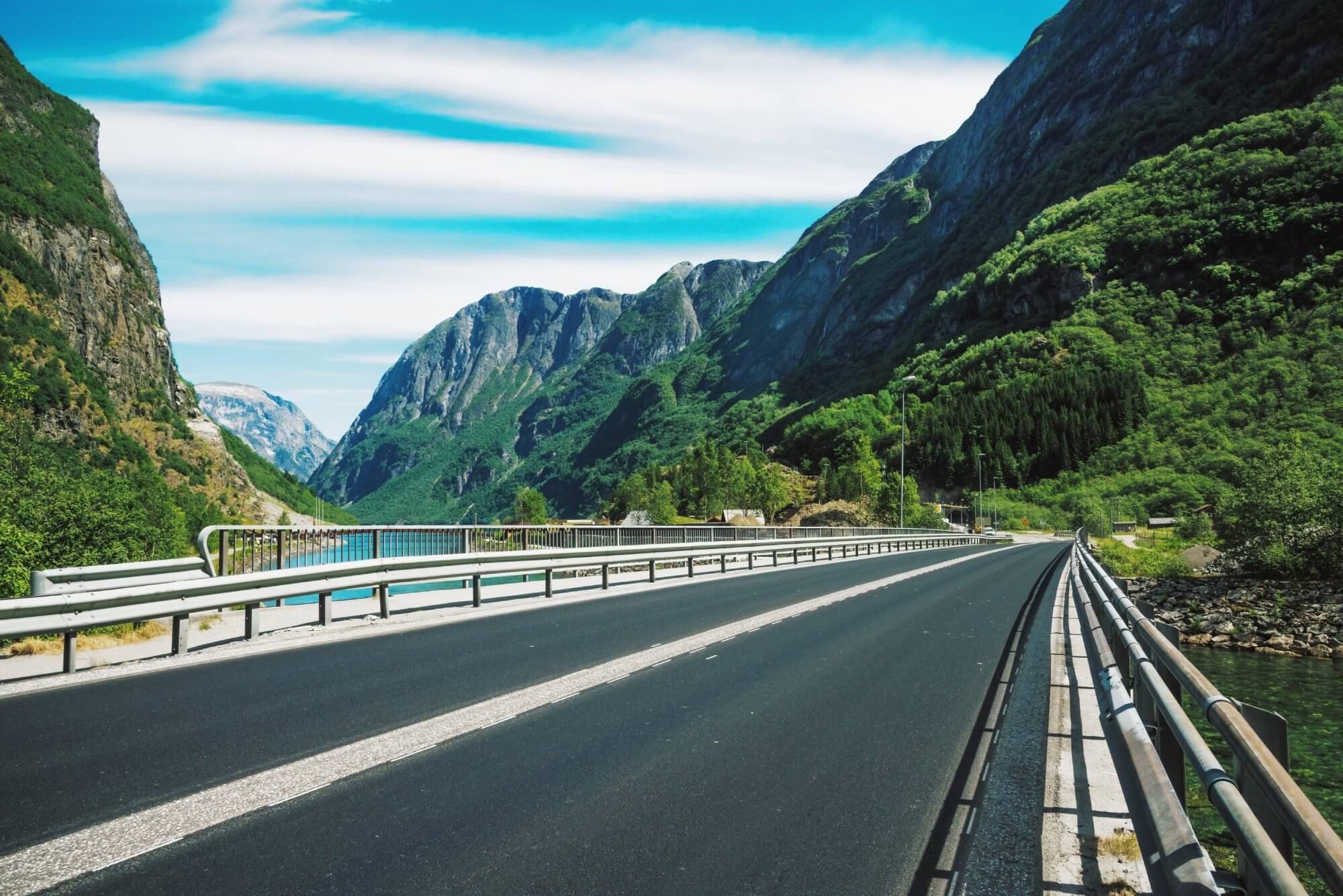 Clear asphalt road with metal guardrails running across a bridge surrounded by green forested mountains under a mostly blue sky with white clouds.