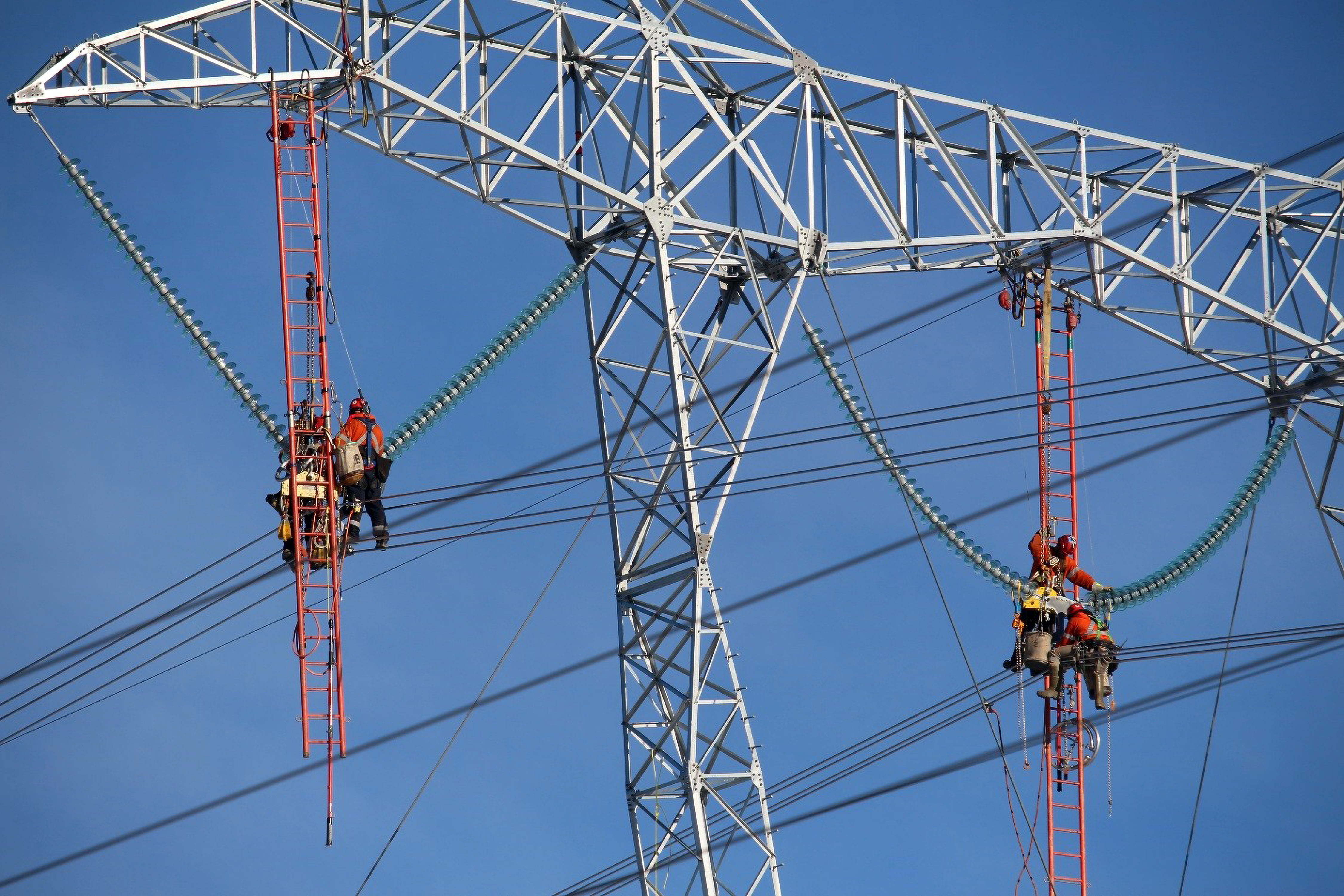 Two lineworkers on a transmission tower with a bird's-eye view of overhead conductors and insulators