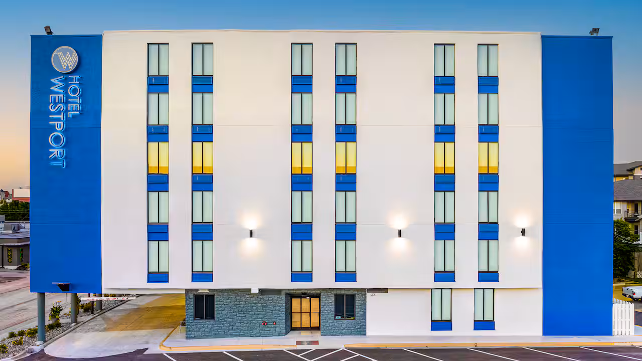 Front view of Hotel Westport building with white and blue exterior, vertical rows of windows, lit wall sconces, and empty parking spaces in front at dusk.