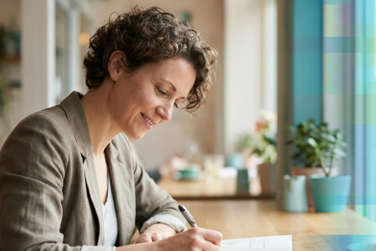 A smiling woman writing notes in a notebook at a cafe table