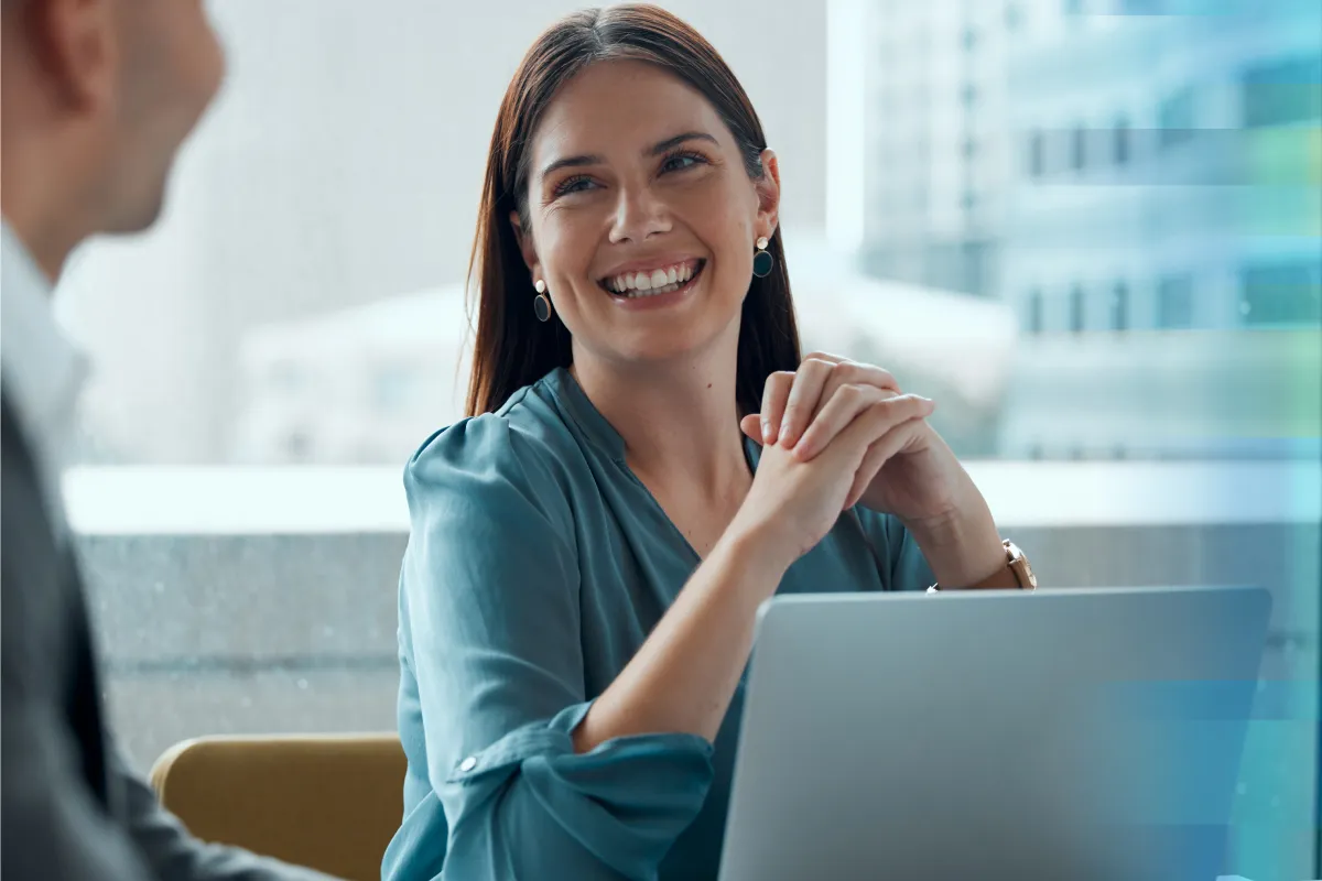Smiling woman in a teal blouse laughing with a colleague near a laptop in a bright office