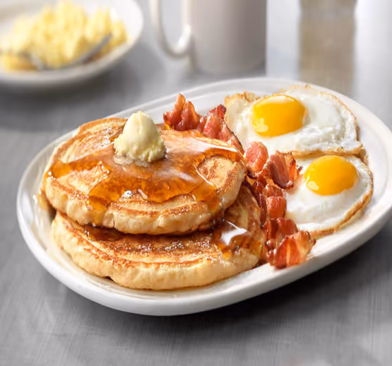 Plate with two pancakes topped with butter and syrup, two sunny-side-up eggs, and crispy bacon strips on the side, set on a table.