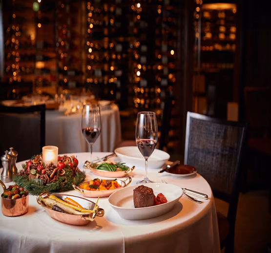 Elegant dining table set with a white tablecloth, featuring a plated steak with raspberry garnish, dishes of roasted vegetables and green beans, two glasses of red wine, a lit candle centerpiece with pine cones and red berries, and a wine cellar backdrop.