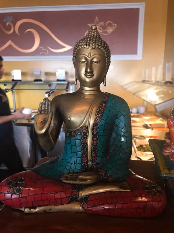 Decorative Buddha statue with a raised right hand in a gesture and left hand resting on the lap, wearing a mosaic robe with turquoise and red tiles, seated in a cross-legged position on a wooden surface, with a blurred background of a buffet setting and a person in motion.