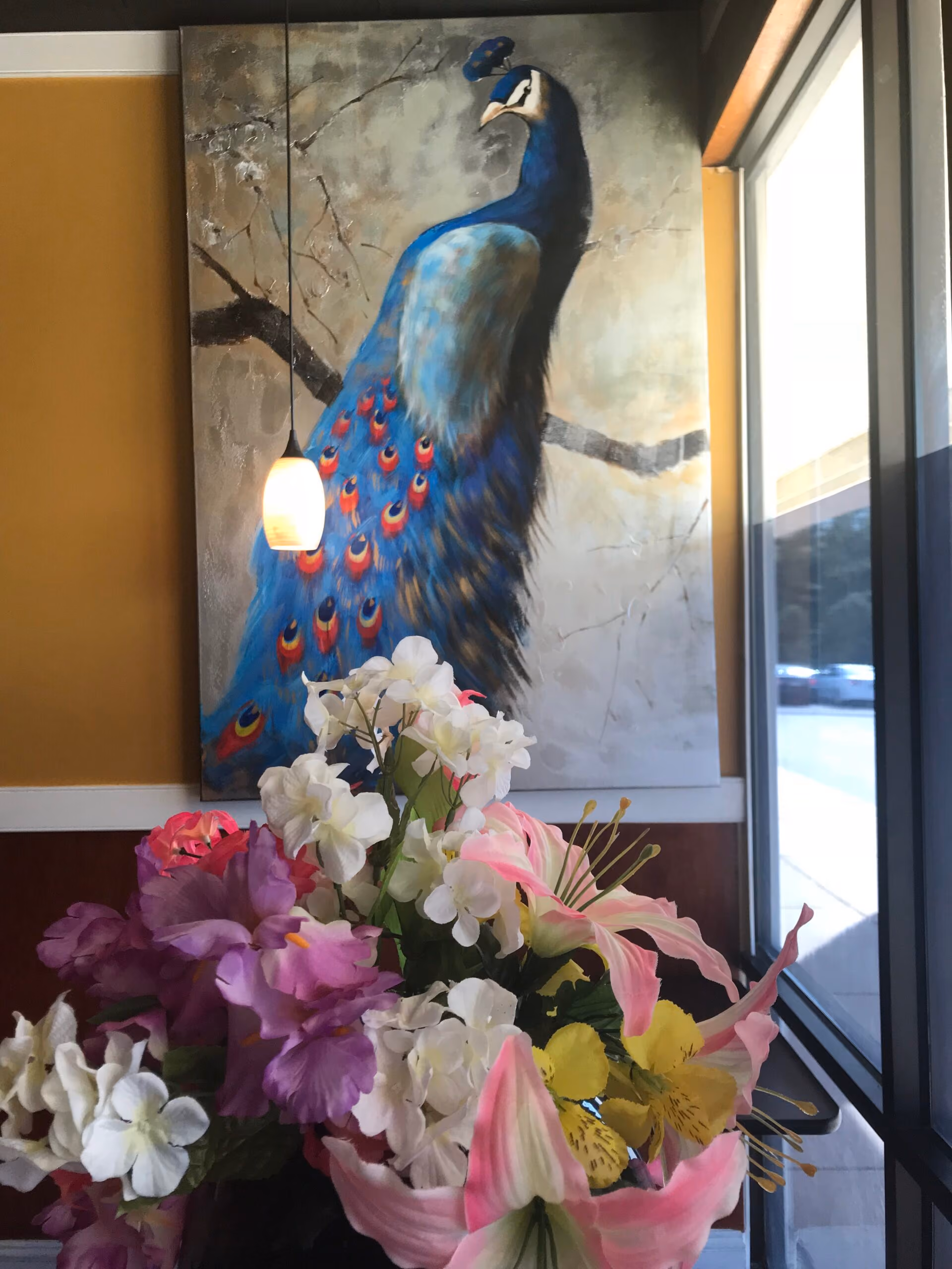 Interior view of a room with a large peacock painting on the wall and a colorful bouquet of flowers in the foreground near a window.