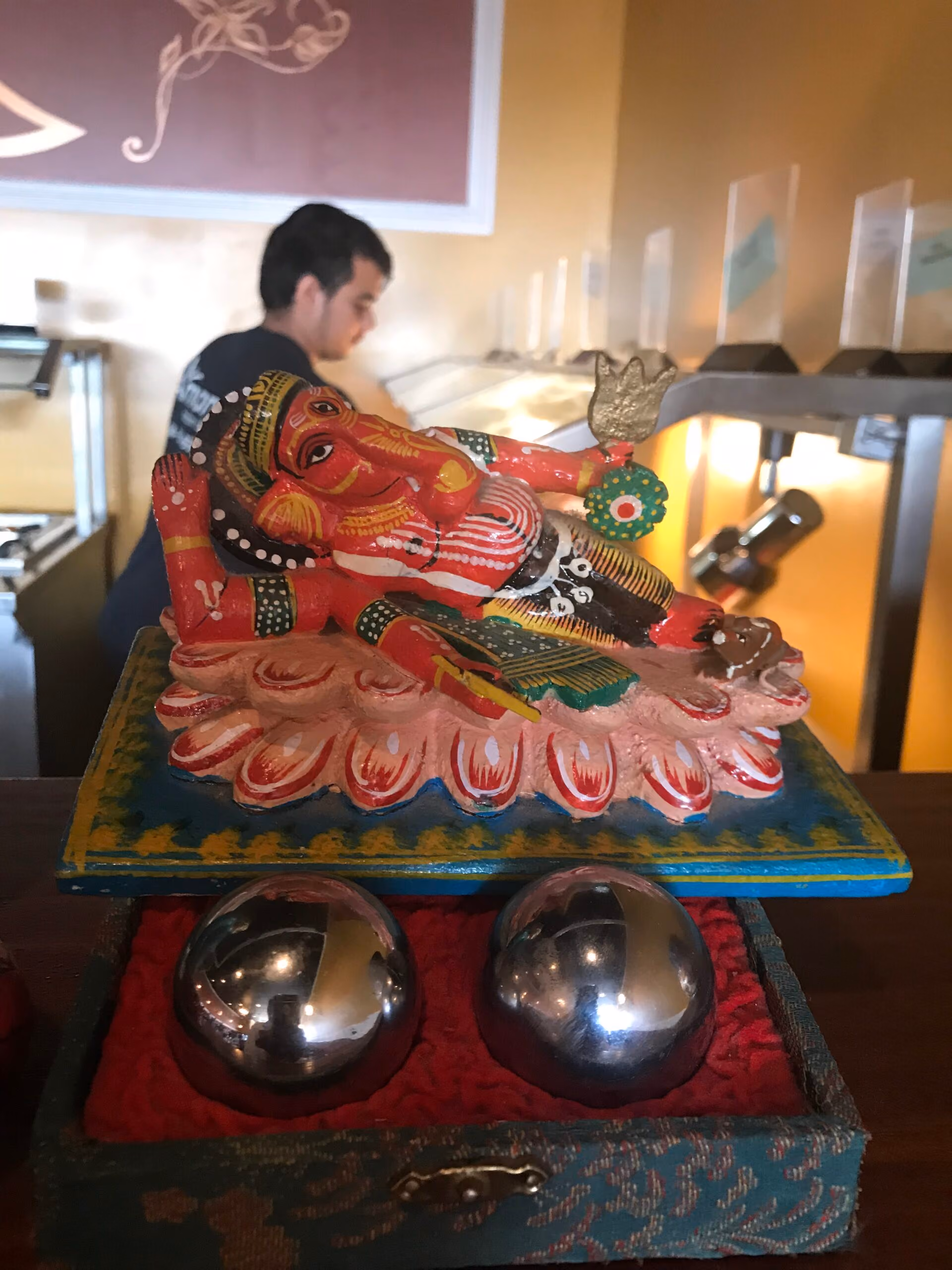 A colorful painted statue of the Hindu god Ganesha reclining on a decorative base, with two silver metallic balls placed below in a cushioned box, and a blurred person and interior background behind.