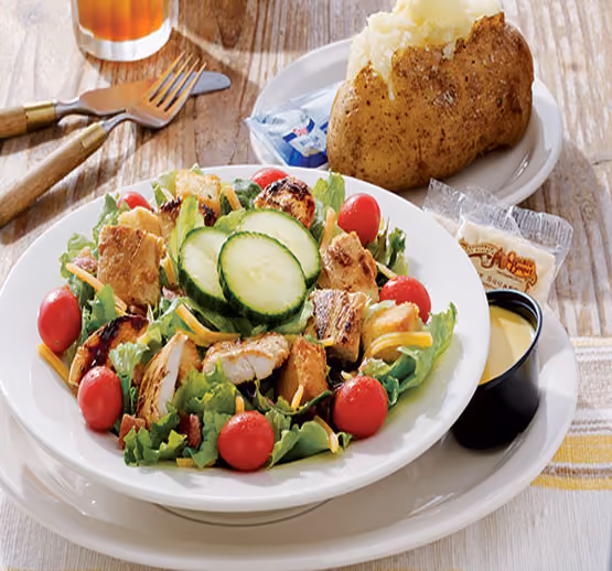 Plate of grilled chicken salad with lettuce, cherry tomatoes, cucumber slices, and shredded cheese, accompanied by a baked potato with sour cream packet and a side of dressing in a black cup on a white plate, with fork and knife on a wooden table.