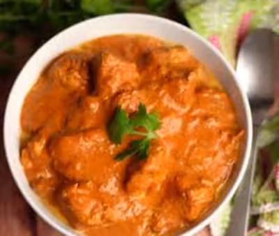 White bowl filled with creamy orange curry garnished with a sprig of cilantro, placed on a wooden surface with a spoon and a green napkin nearby.